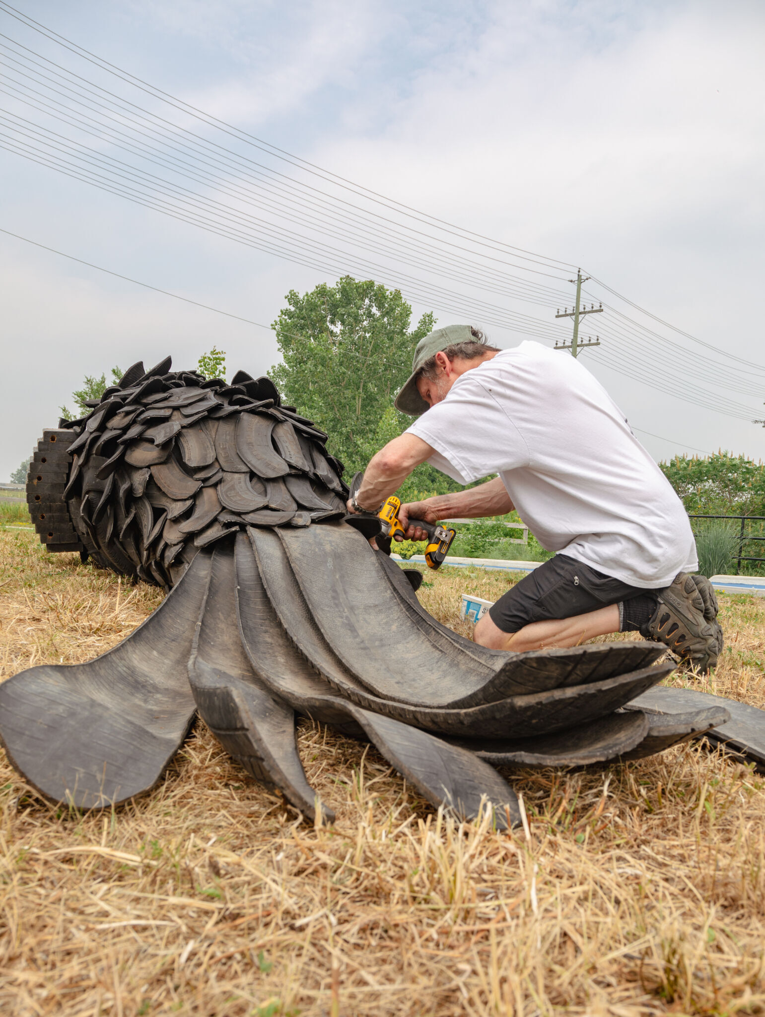 Artiste à genoux qui installe des pièces sur sa sculpture, un corbeau en pneus recyclés de 16 pieds. Vue de dos du corbeau étendu au sol.
