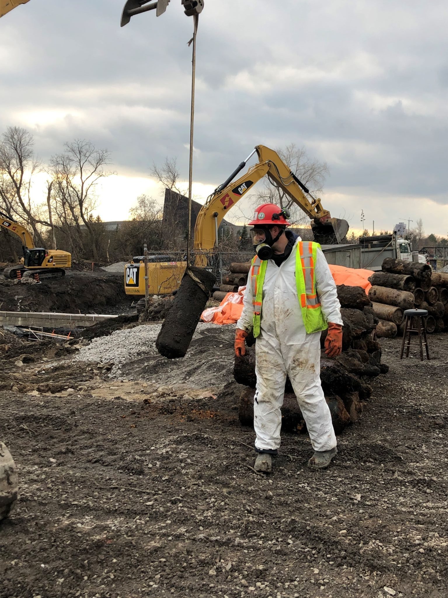 A worker geared up in a carbon-lined suit and full-face respirator stands in front of a pile of acetylene gas cylinders. In the background, there are mechanical excavators.