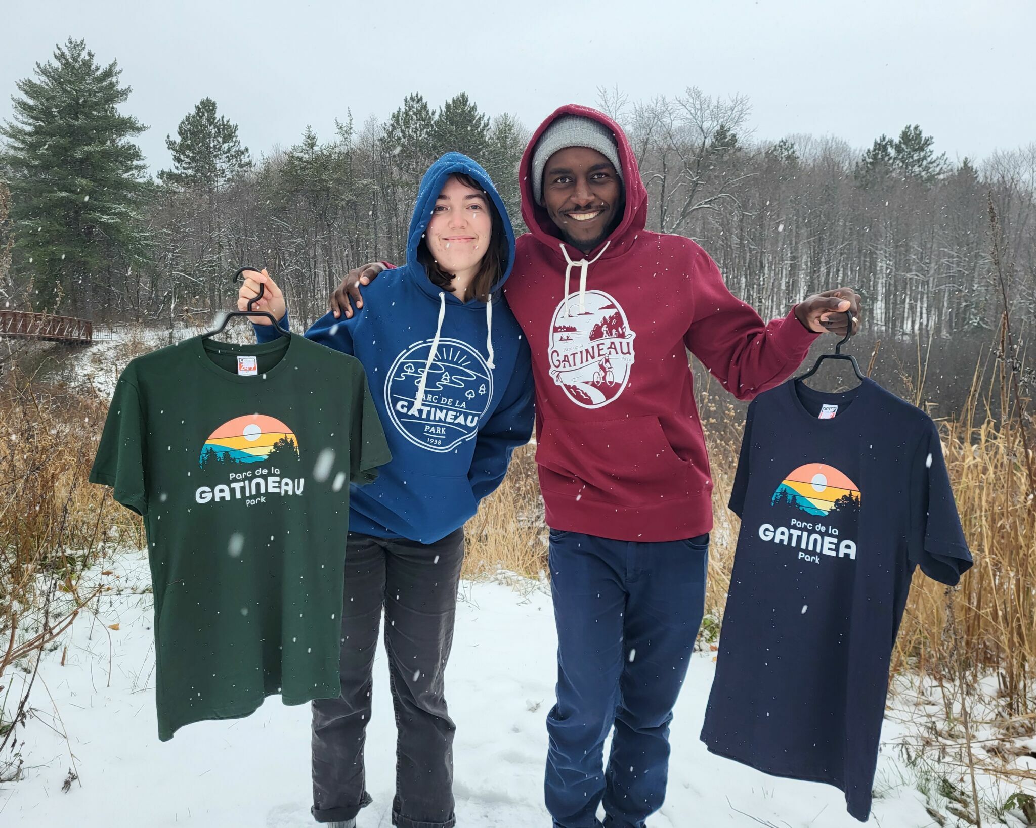 Two people outside in a snowy landscape wearing Gatineau Park clothing and each holding a hanger with Gatineau Park t-shirts.