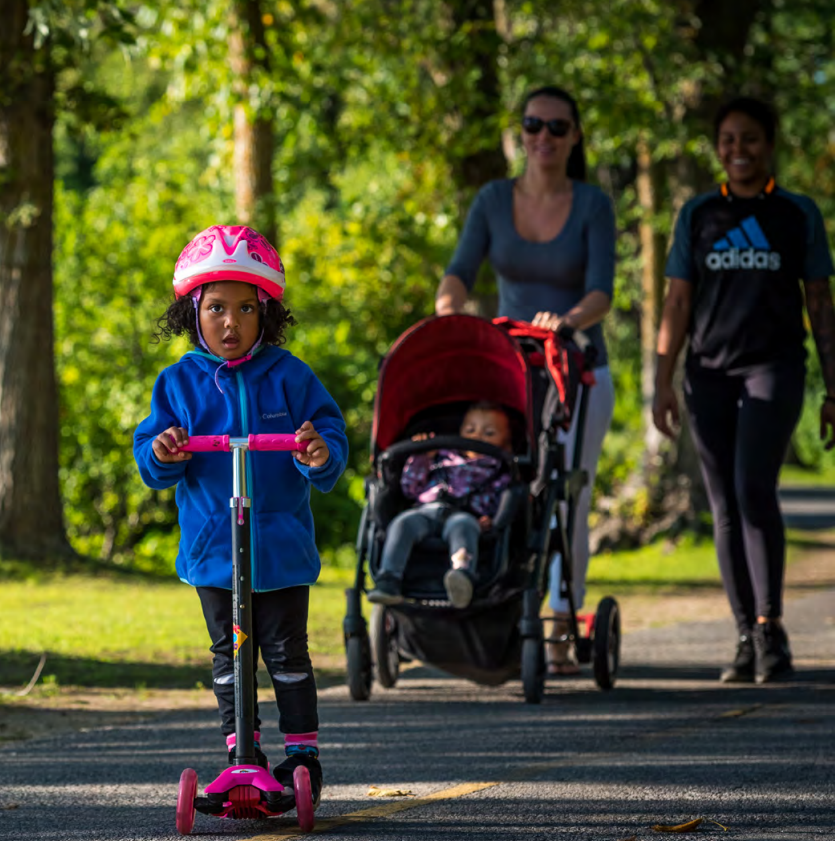 Two people walking side-by-side on the Capital Pathway with a stroller, behind a child on a scooter.