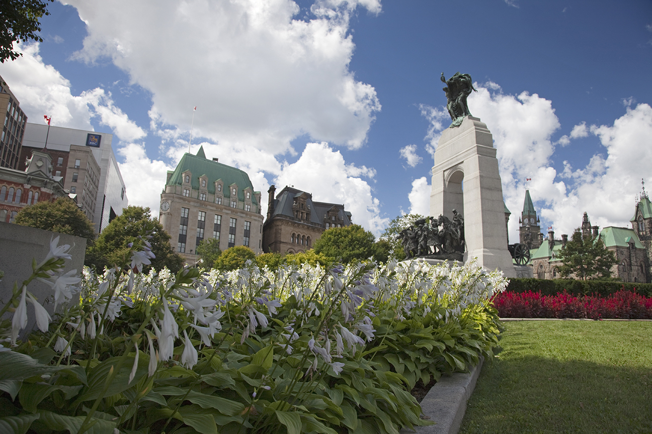 Vue du Monument commémoratif de guerre, des édifices du Parlement et du bureau de poste; des parterres de fleurs sont à l’avant-plan.