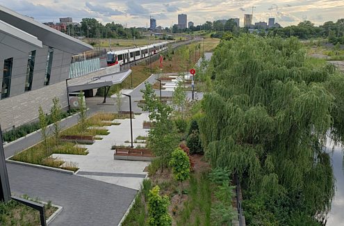 Vue, vers l’ouest, de l’esplanade de la station Pimisi de l’O-Train, avec ses bancs, son aménagement paysager et ses supports à vélos. Le tronçon 1 de l’éventuel sentier des Plaines-LeBreton se poursuivra à l’ouest, au bout de l’esplanade.