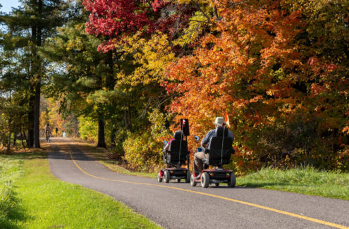 Two people riding four-wheel electric scooters on a multi-use pathway, and a cyclist riding in the opposite direction, in the fall