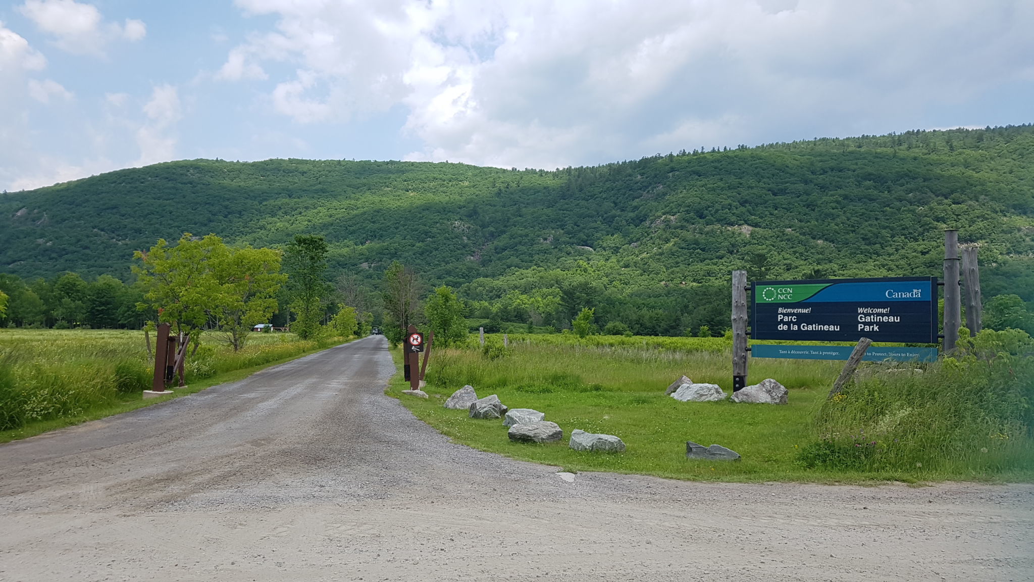 The Luskville Falls area in summer. Welcome sign and gravel road in the foreground. The hills of the Outaouais in the background.