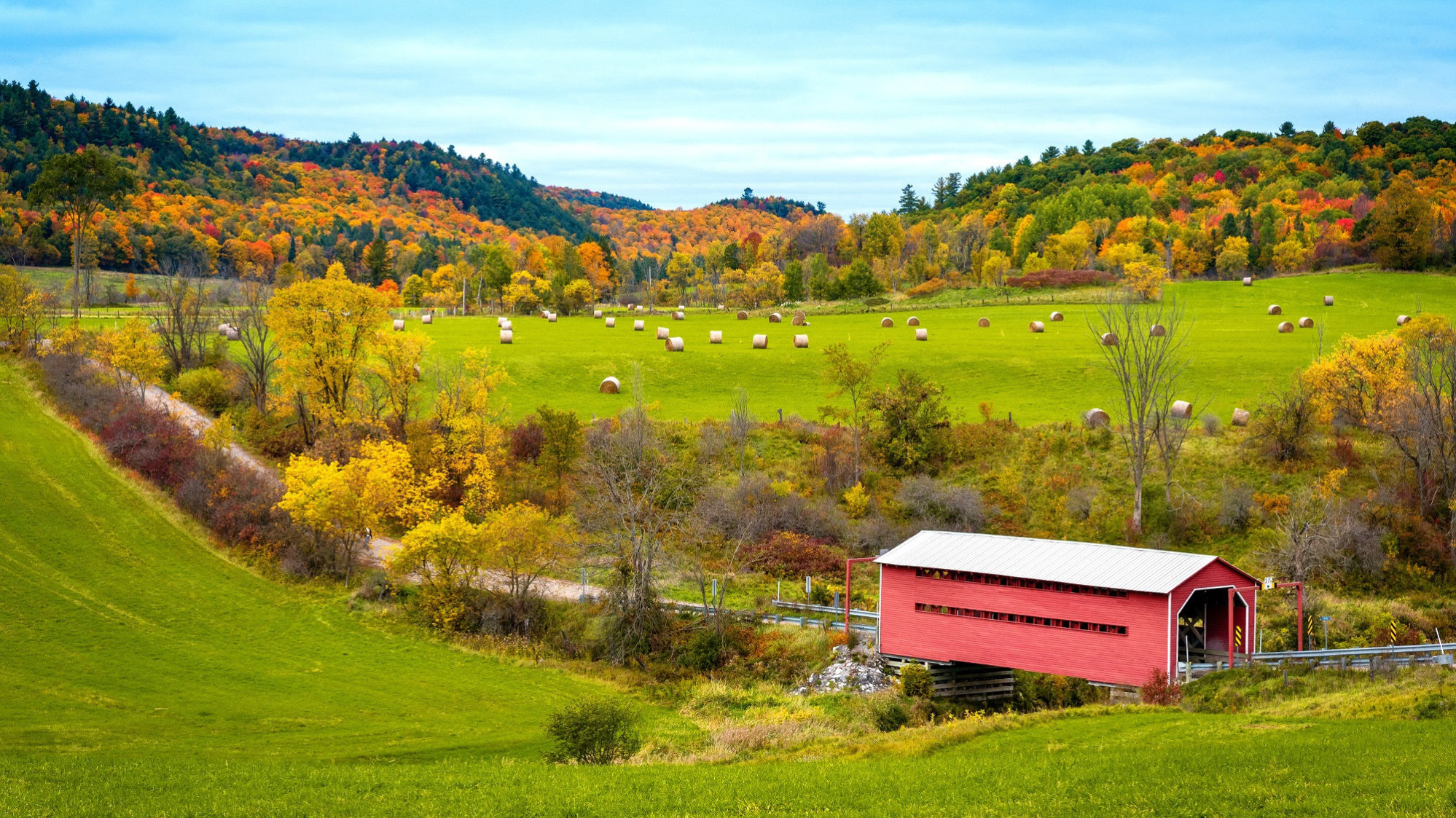 View of Meech Creek Valley in the fall. Scene of a covered bridge and haybales, with ochre- and orange-coloured trees in the background.